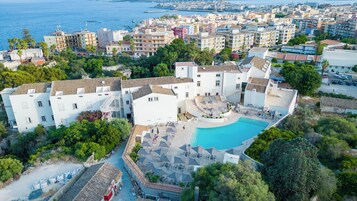 A coastal hotel with a swimming pool, multiple balconies, and a view of the sea.