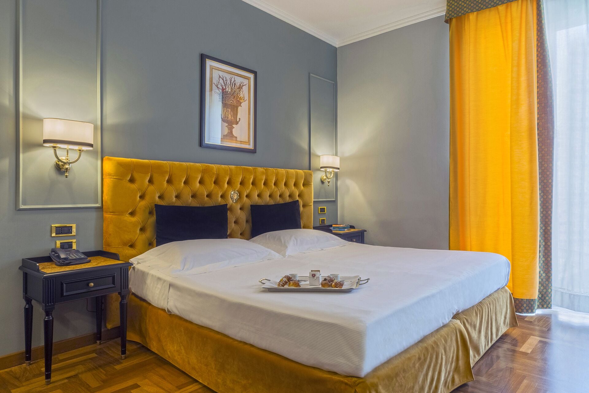 Une chambre d’hôtel avec un grand lit, deux tables de chevet, une petite table, un tableau encadré au mur et une fenêtre avec des rideaux jaunes.
