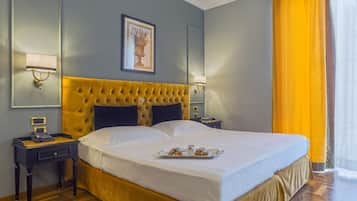 Une chambre d’hôtel avec un grand lit, deux tables de chevet, une petite table, un tableau encadré au mur et une fenêtre avec des rideaux jaunes.