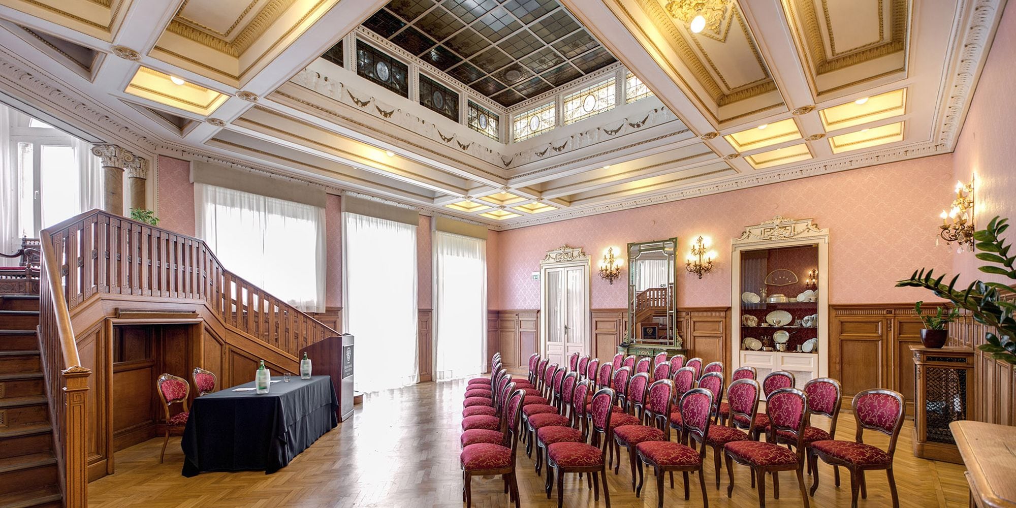 A grand hall with a high ceiling, ornate columns, and a staircase leading to an upper level. Rows of chairs are arranged for seating.