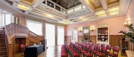 A grand hall with a high ceiling, ornate columns, and a staircase leading to an upper level. Rows of chairs are arranged for seating.