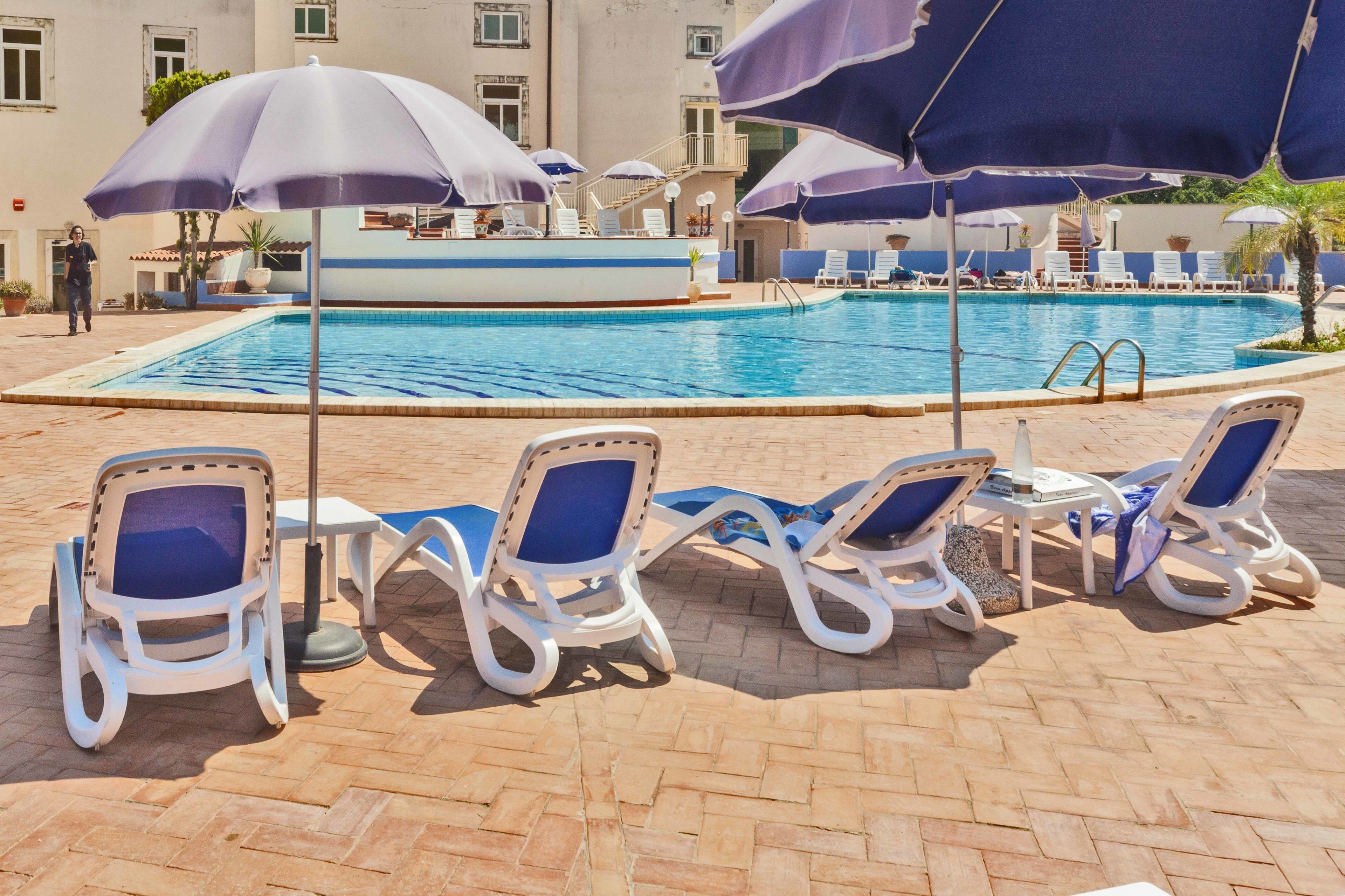 A poolside area with lounge chairs and umbrellas, a swimming pool, and a building in the background.