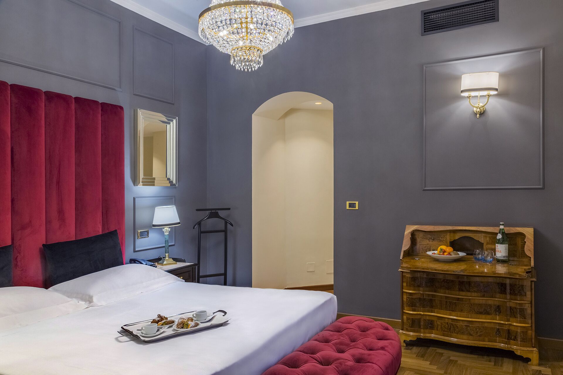 A hotel room with a large bed, a red upholstered headboard, a chandelier, a wall-mounted lamp, a wooden dresser, and a mirror.