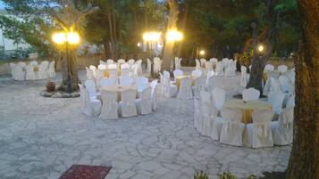An outdoor event space with round tables and chairs arranged in a semi-circle, surrounded by trees and stone flooring.