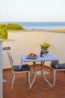 A terrace with a table set for two, featuring a plate of fruit, a bottle of wine, and a vase of flowers.