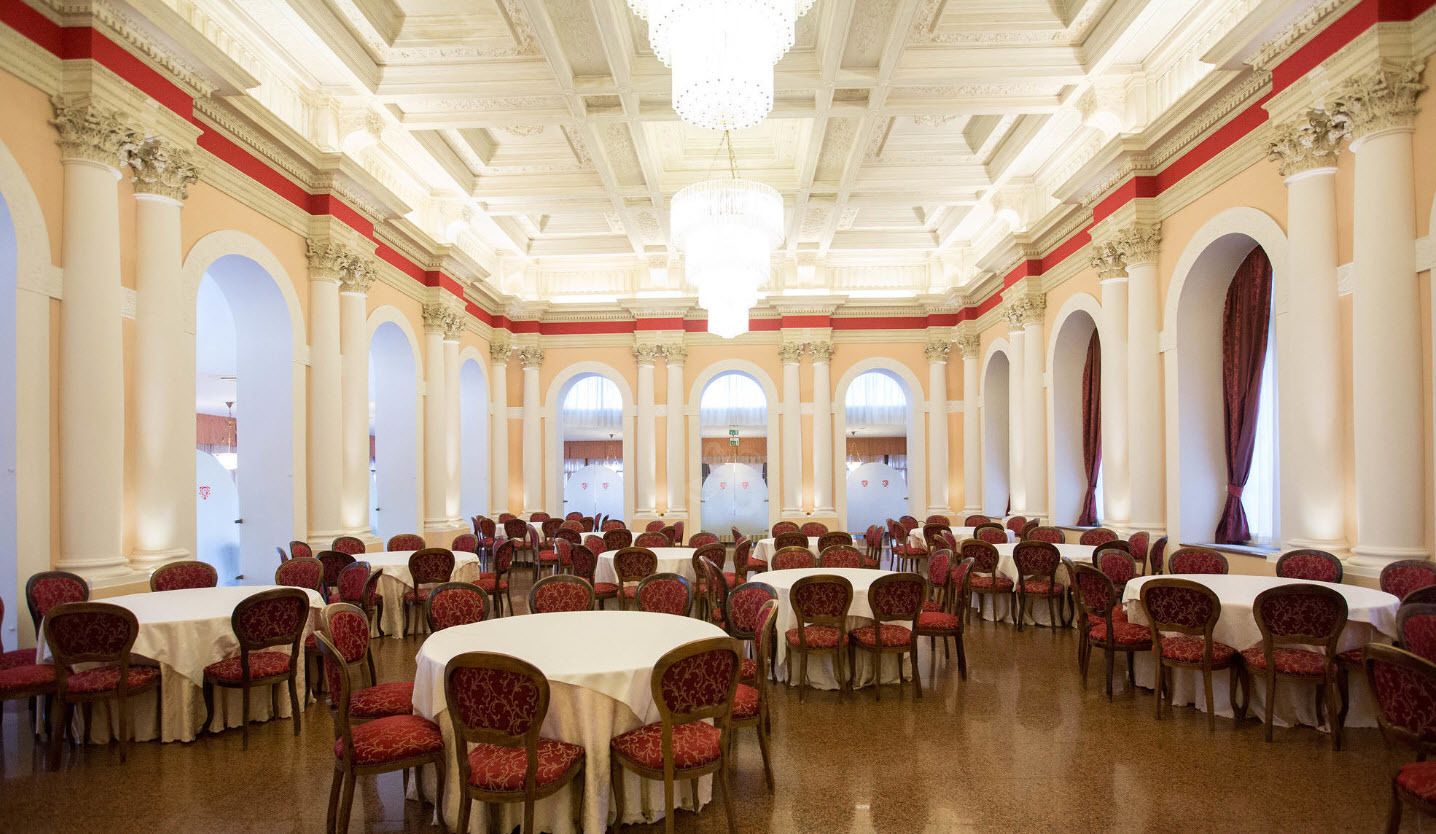 Une vaste salle de banquet avec des tables rondes et des chaises, de hauts plafonds ornés de moulures et de grandes fenêtres avec des rideaux.