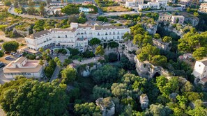 An aerial view of a coastal town with a prominent white building featuring a central courtyard and surrounding greenery. - Grand Hotel Villa Politi (Syracuse)