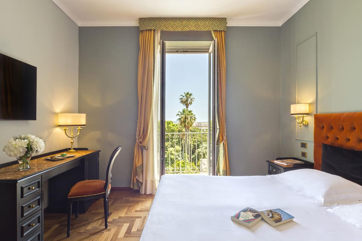 A hotel room with a bed, a desk, a chair, a nightstand, and a window with curtains.