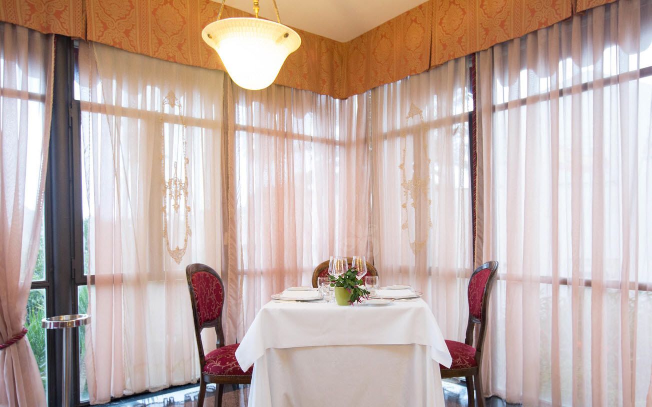 A dining room with a table set for two, large windows with sheer curtains, and a chandelier.