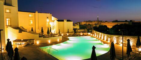 A swimming pool with lounge chairs and umbrellas, surrounded by buildings with a staircase and a view of the city skyline.