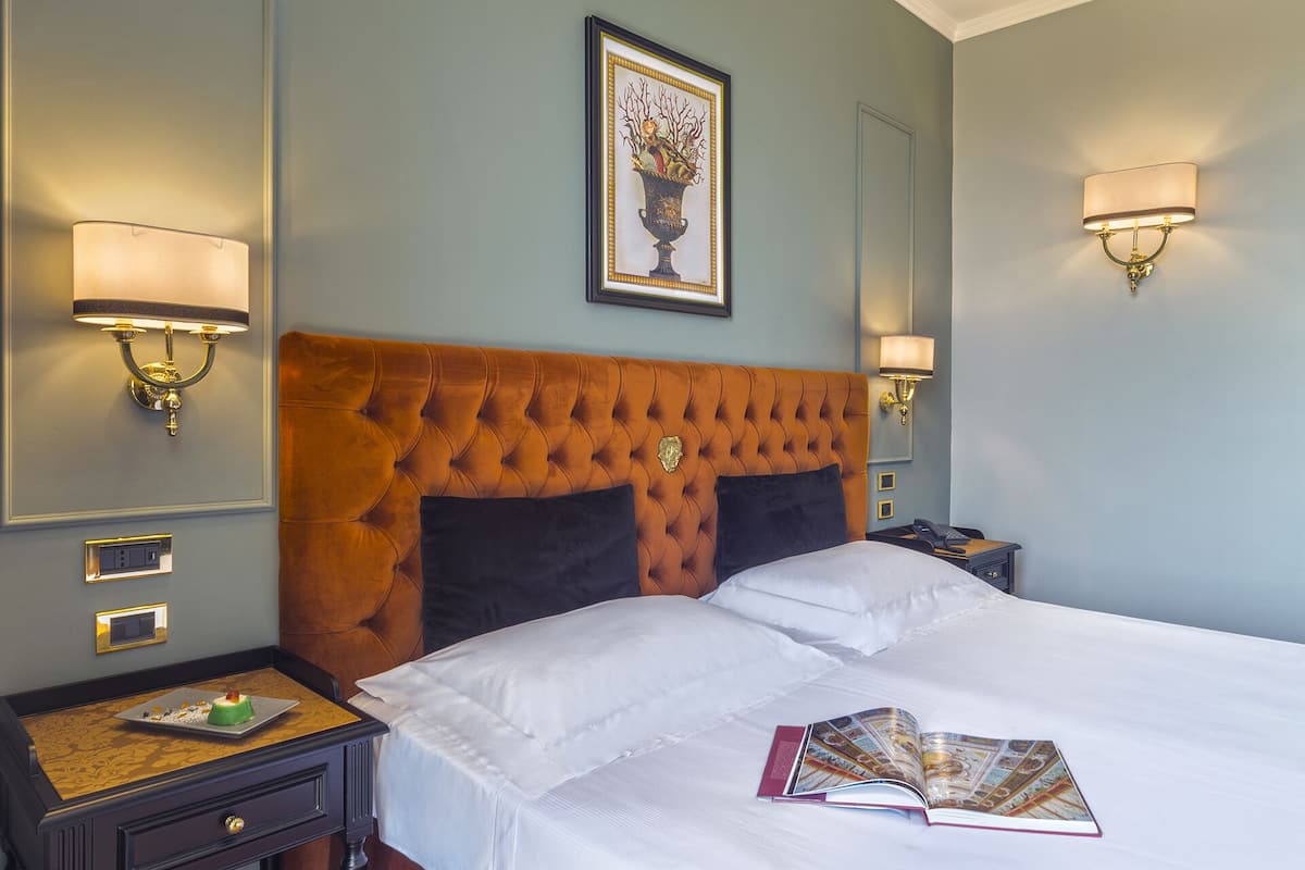 A hotel room with a tufted headboard, a bed with white linens, two bedside tables with lamps, and a framed artwork above the headboard.