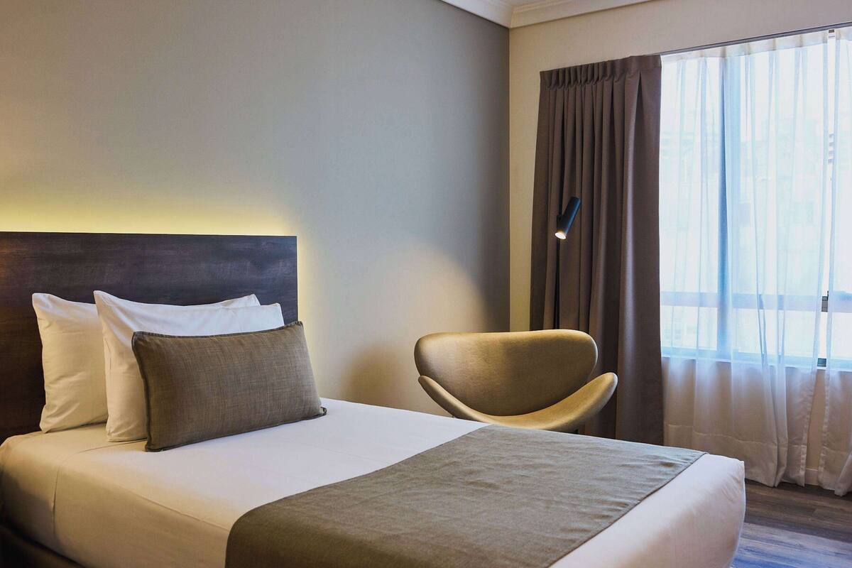 A hotel room with a bed, pillows, a chair, and a window with curtains.
