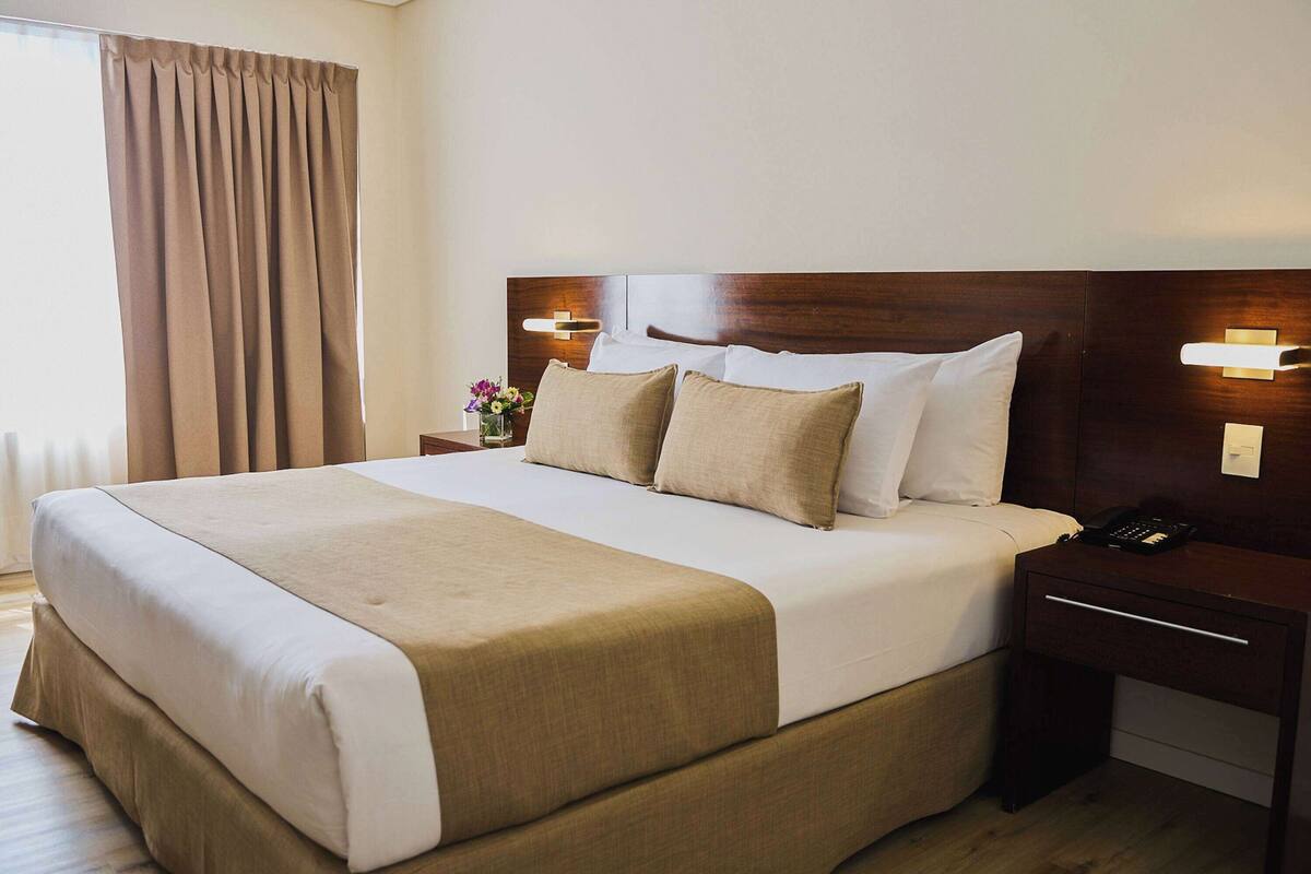 A hotel room with a large bed, a nightstand with a phone, and a wall-mounted light fixture.