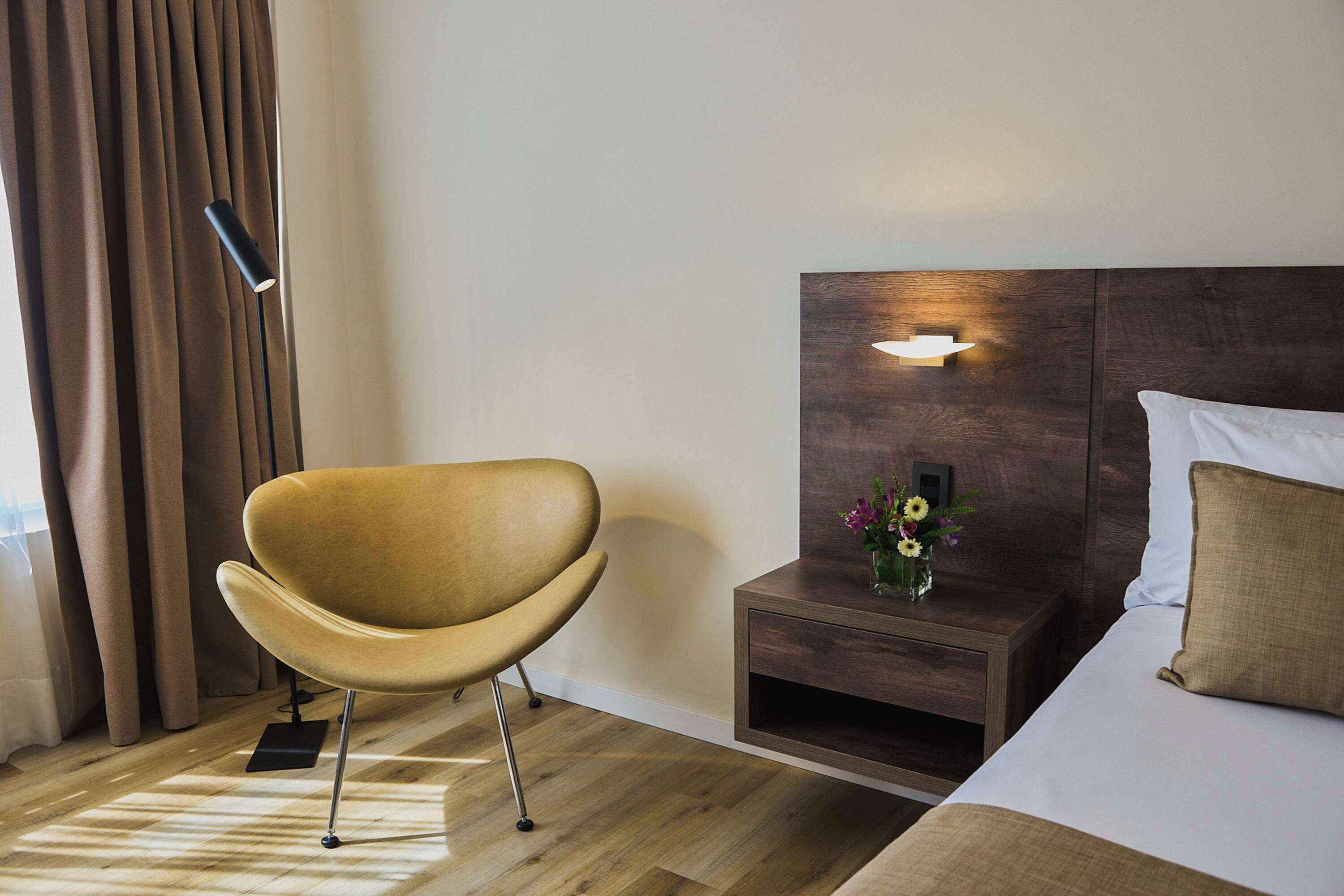 Ein Hotelzimmer mit einem modernen Bett, einem Nachttisch aus Holz mit einer Blumenvase, einem Sessel und einer Stehlampe.