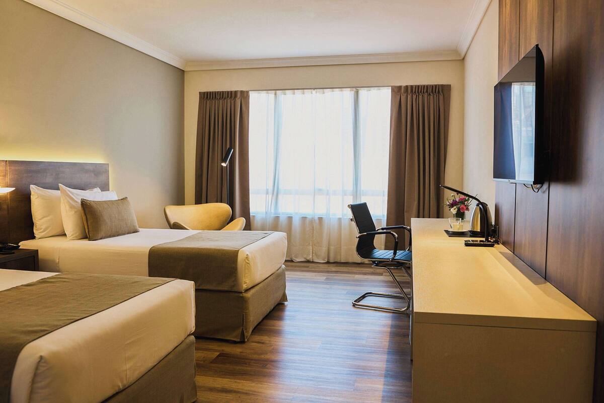 A hotel room with two beds, a desk, a chair, a TV, and a window with curtains.