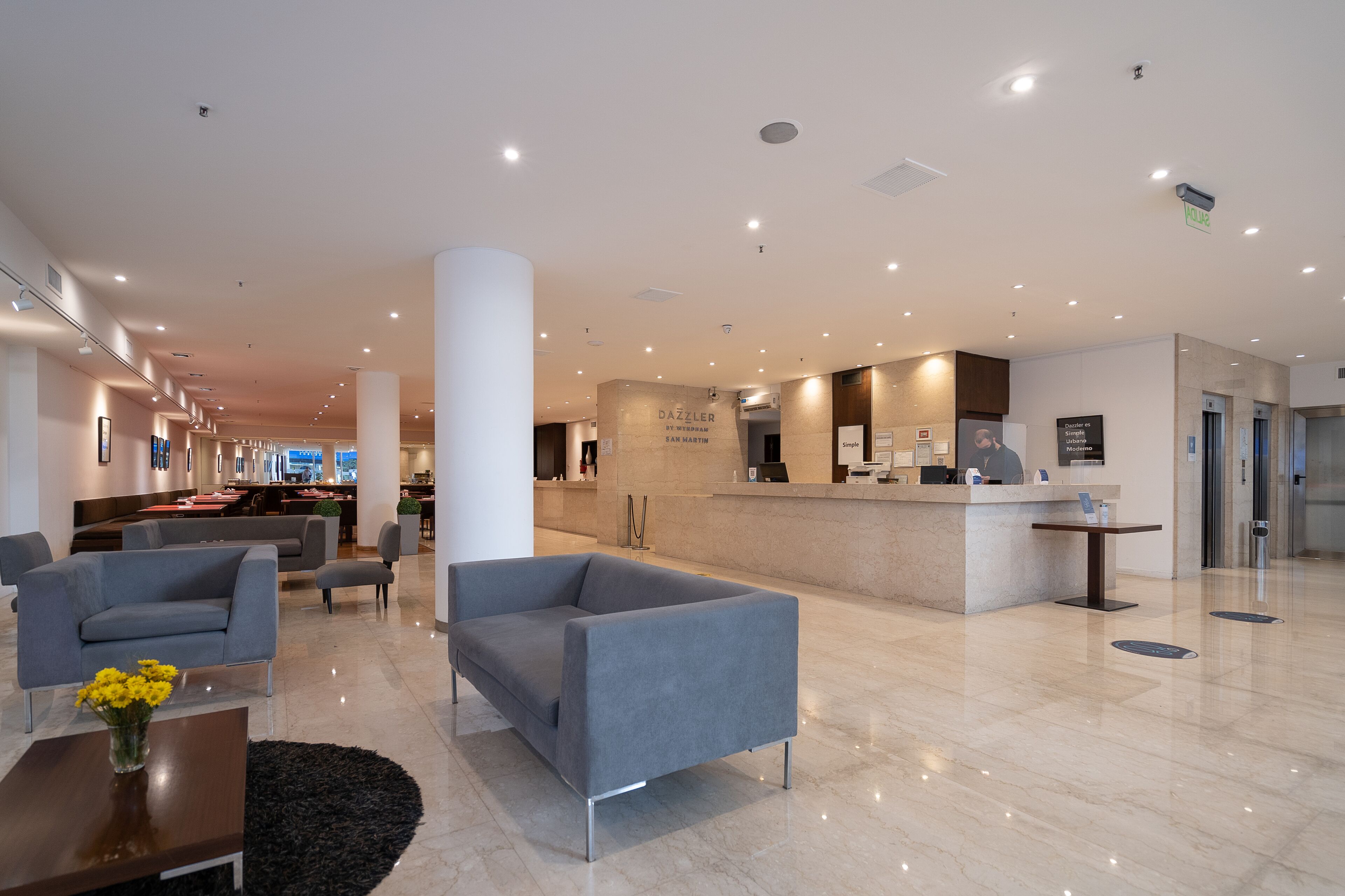 A modern hotel lobby with a reception desk, seating area, and a dining space in the background.