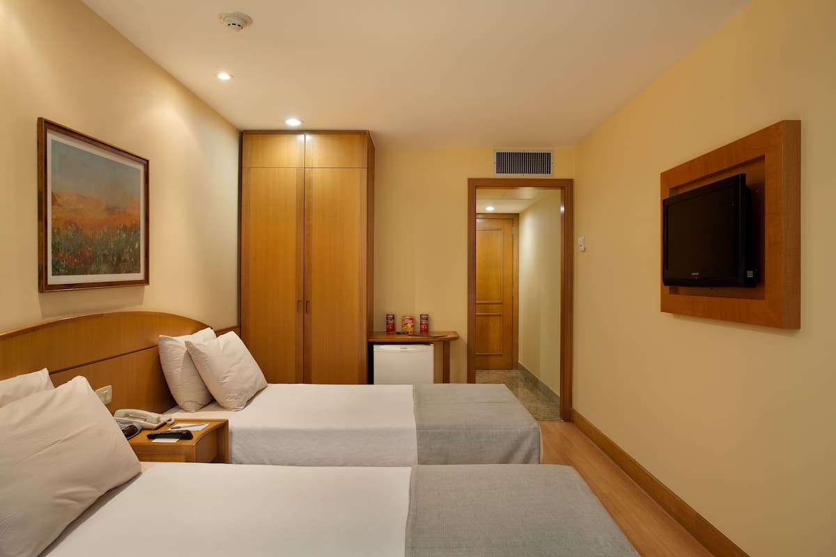 Ein Hotelzimmer mit zwei Betten, einem an der Wand befestigten Fernseher, einem kleinen Tisch mit Telefon und Fernbedienung sowie einem Bild an der Wand.