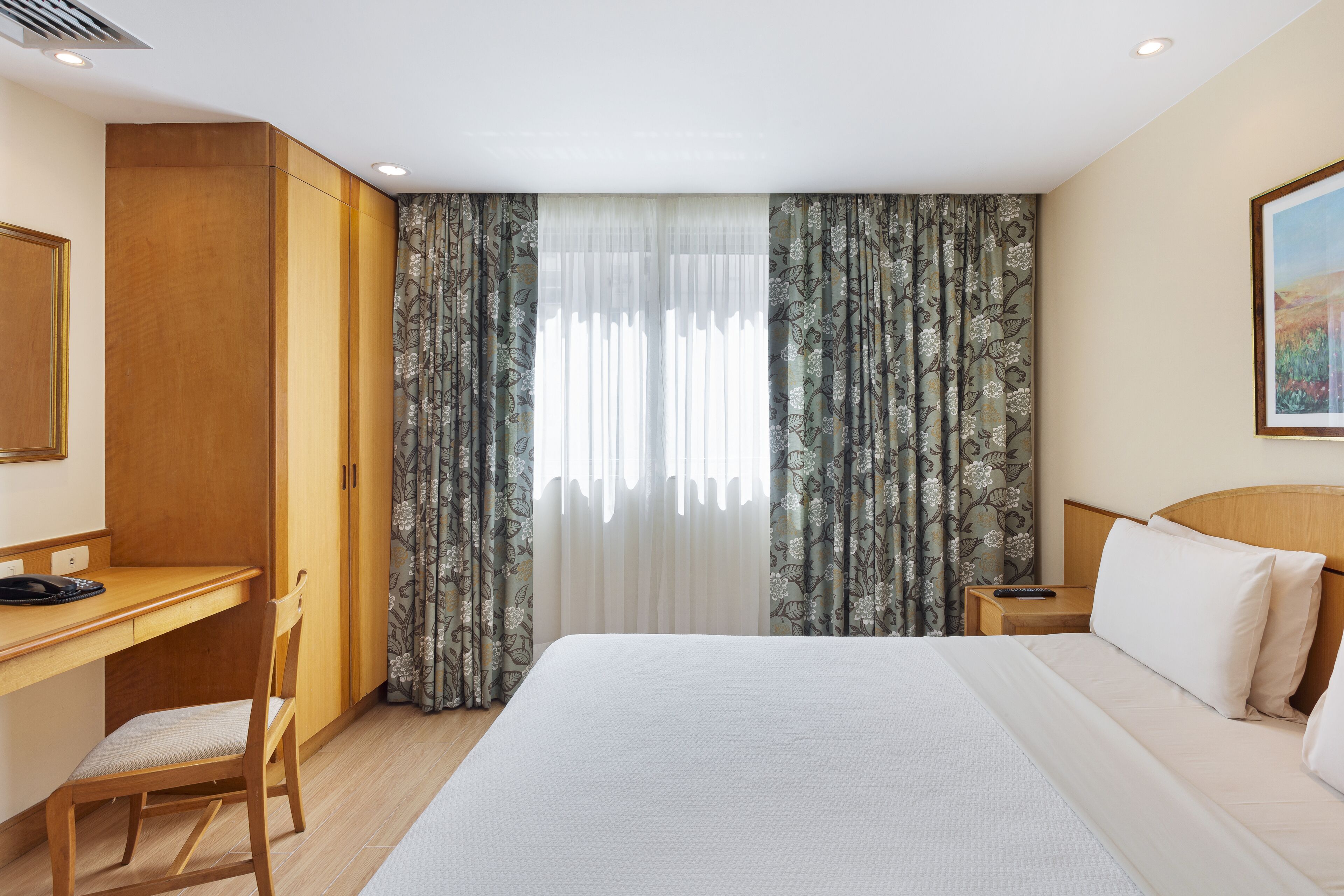 A hotel room with a bed, a desk, a chair, a nightstand, and curtains.