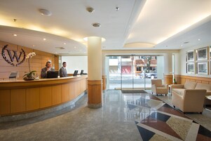 A hotel reception area with a wooden reception desk, two receptionists, a large pillar, and a patterned carpet.