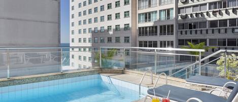 Una piscina en la azotea con sillas de descanso y barandilla de cristal, rodeada por un edificio de gran altura.