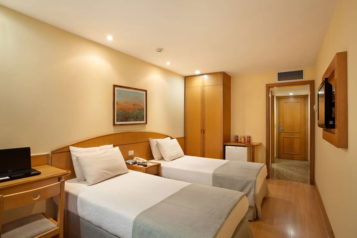 Ein Hotelzimmer mit zwei Betten, einem Schreibtisch mit einem Laptop, einem Bild an der Wand, einem Kleiderschrank, einer Minikühlschrank und einer Tür zu einem weiteren Raum.