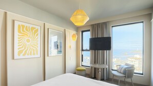 A hotel room with a bed, a TV, a large window with a city view, and artwork on the wall.
