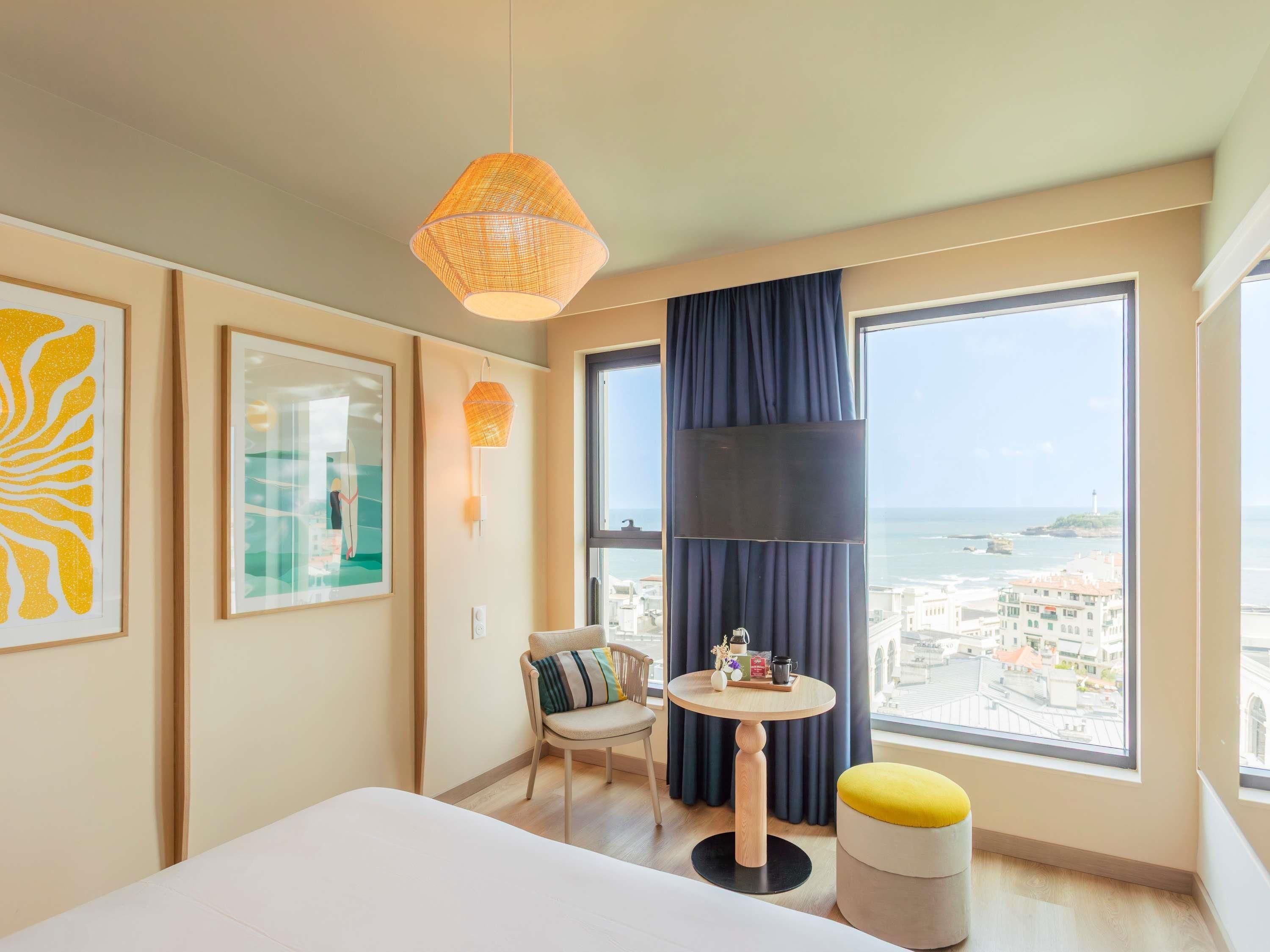 Une chambre d’hôtel avec un lit, une table ronde et une chaise, un petit tabouret, une grande fenêtre donnant sur la mer et des œuvres d’art accrochées aux murs.