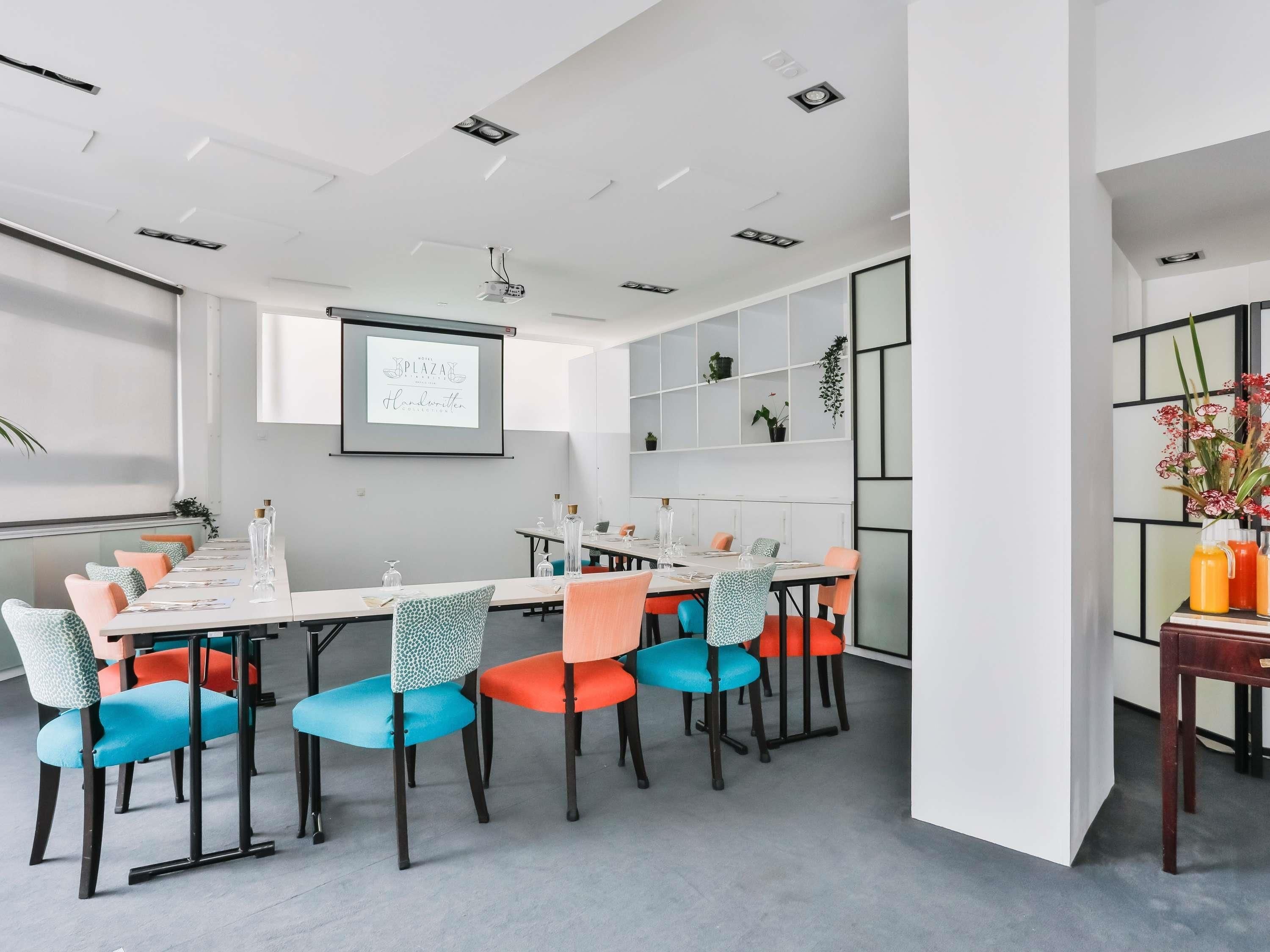 A modern meeting room with a long table, colorful chairs, a projector screen, and a whiteboard.