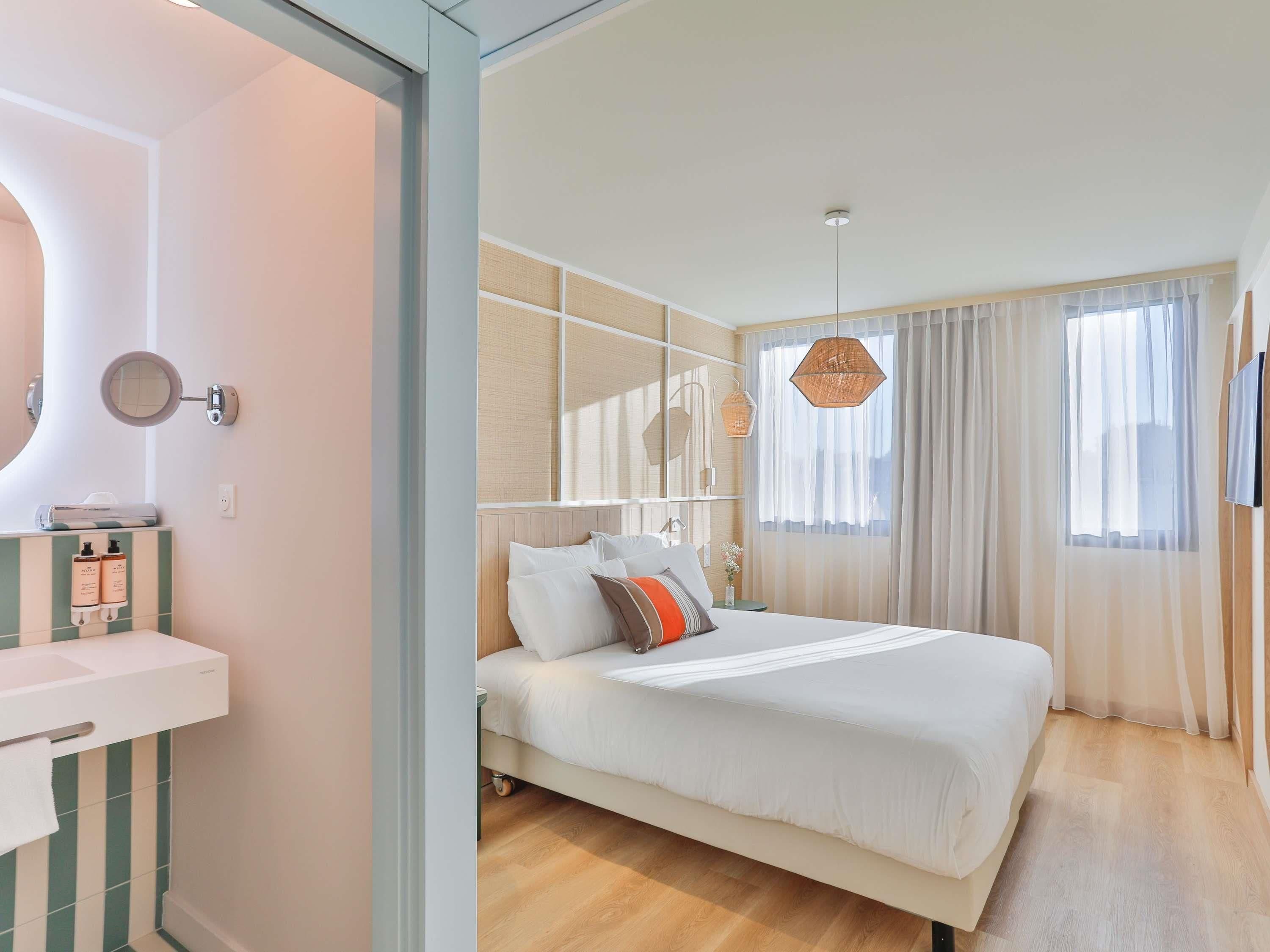 A hotel room with a bed, a bedside table, a mirror, a window with curtains, and a wall-mounted light fixture.