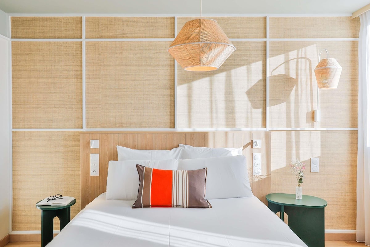 Quarto de hotel moderno com uma cama grande, uma mesa de cabeceira e um criado-mudo verde, com uma luminária fixada na parede.