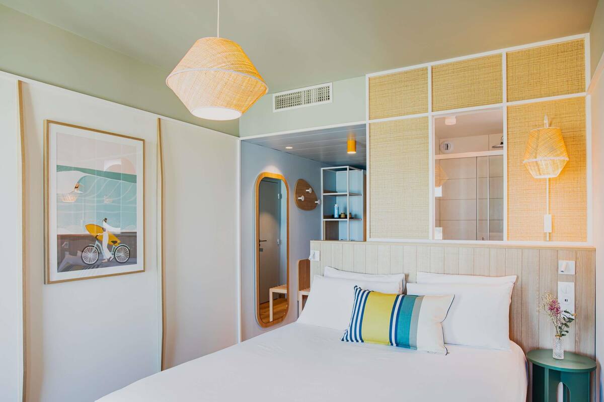 A bedroom with a large bed, a framed picture on the wall, a mirror, and a bedside table with a vase.