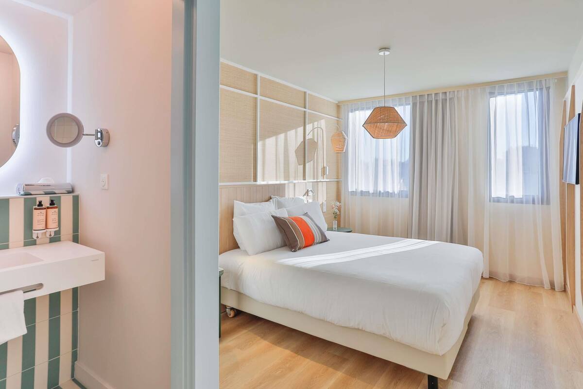 A hotel room with a bed, a bedside table, a mirror, and a window with curtains.