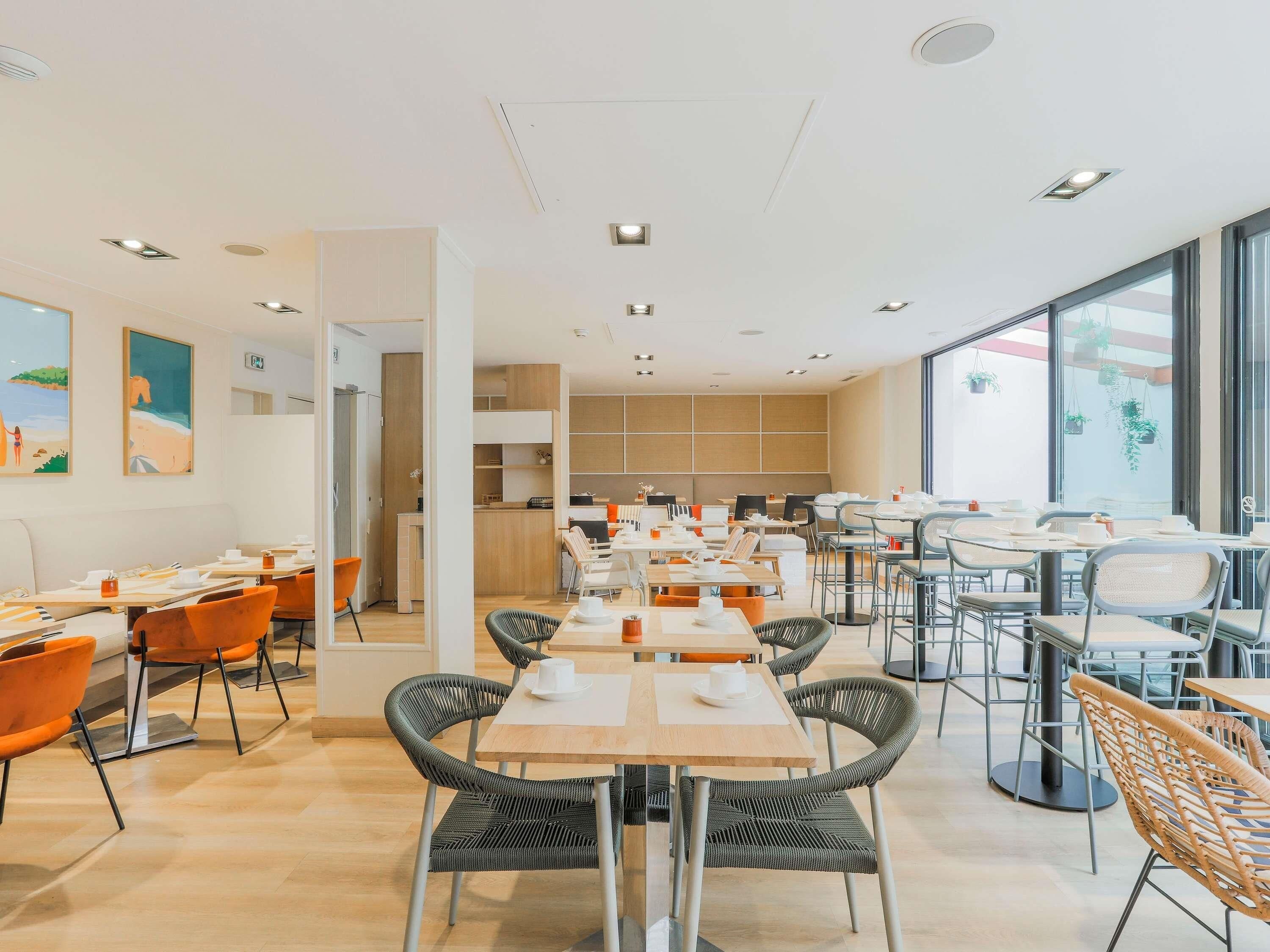 A modern dining area with multiple tables and chairs, a kitchen in the background, and large windows providing natural light.