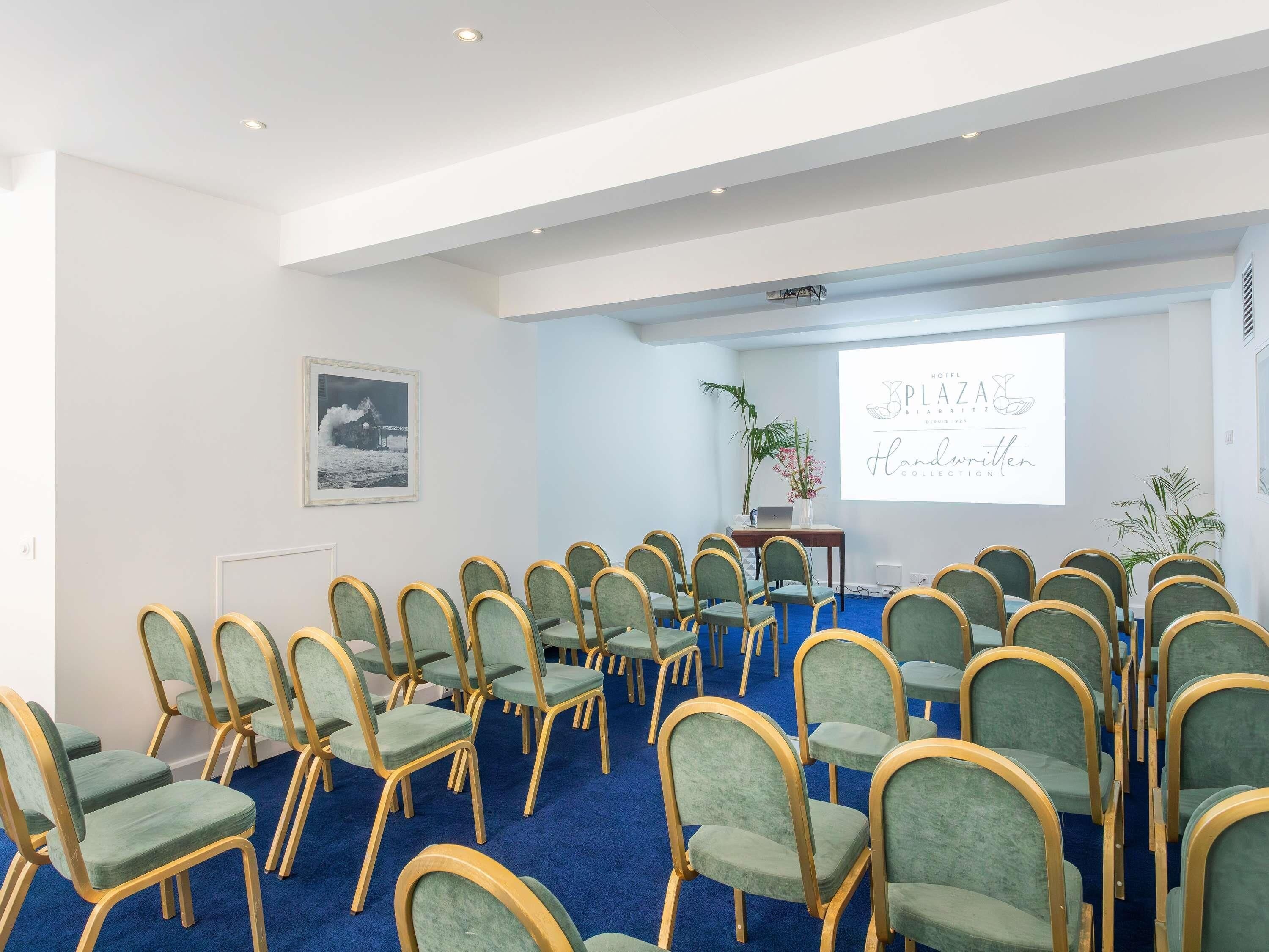 A conference room with rows of green chairs, a projector screen, and a framed picture on the wall.