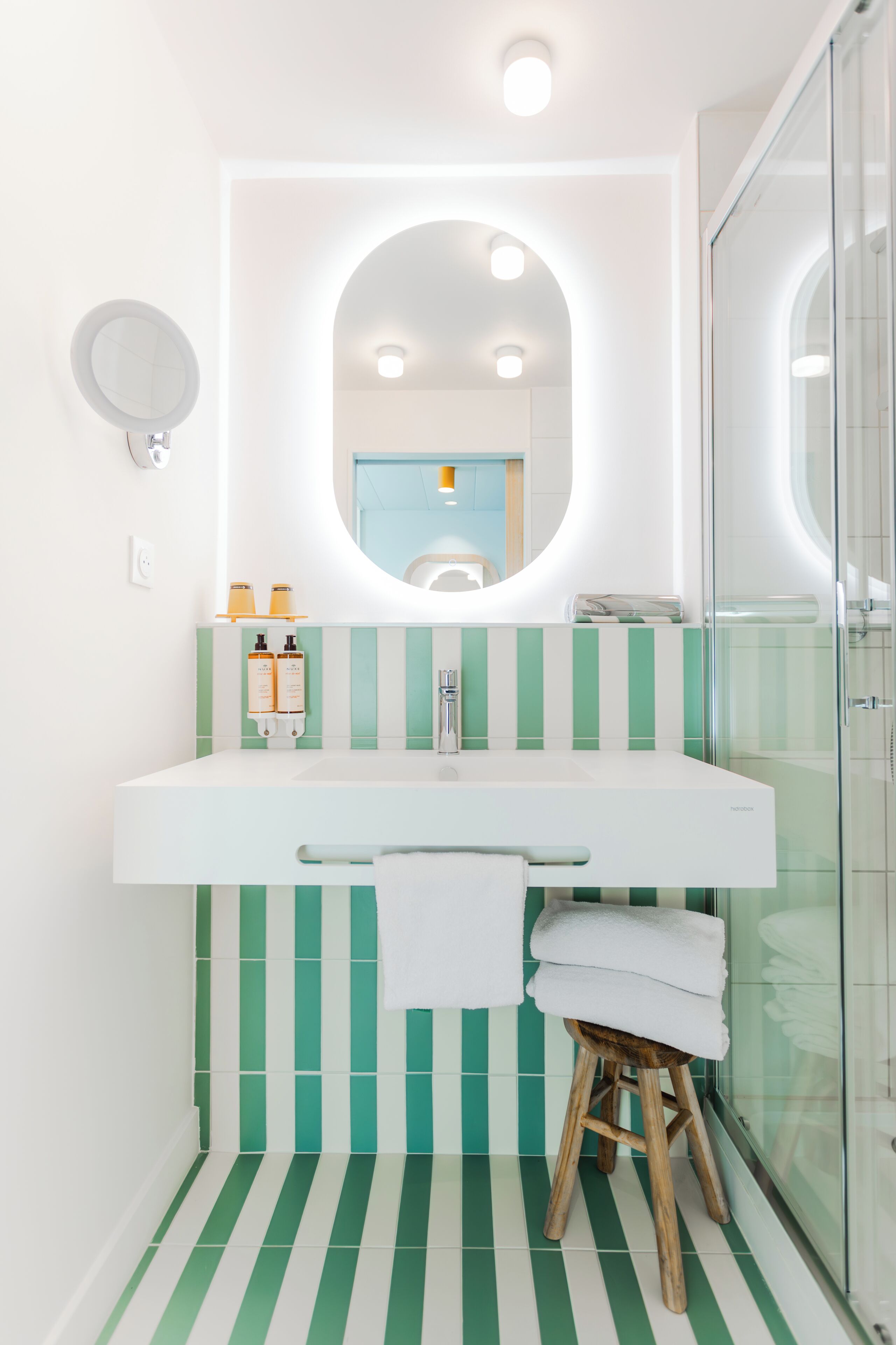 A bathroom with a white sink, green-striped tiles, a wooden stool with a white towel, and a glass shower enclosure.