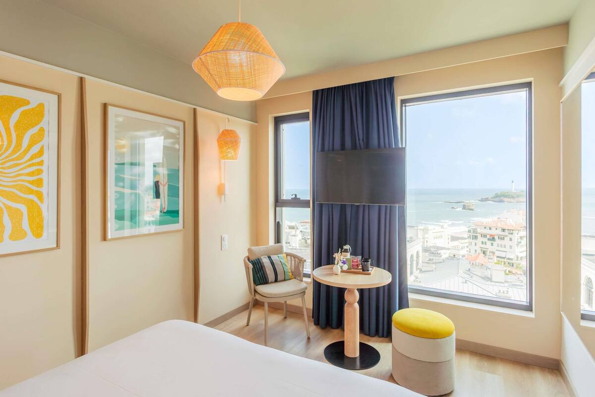 A hotel room with a bed, a round table with a chair, a small stool, a large window with a view of the sea, and two wall-mounted lamps.