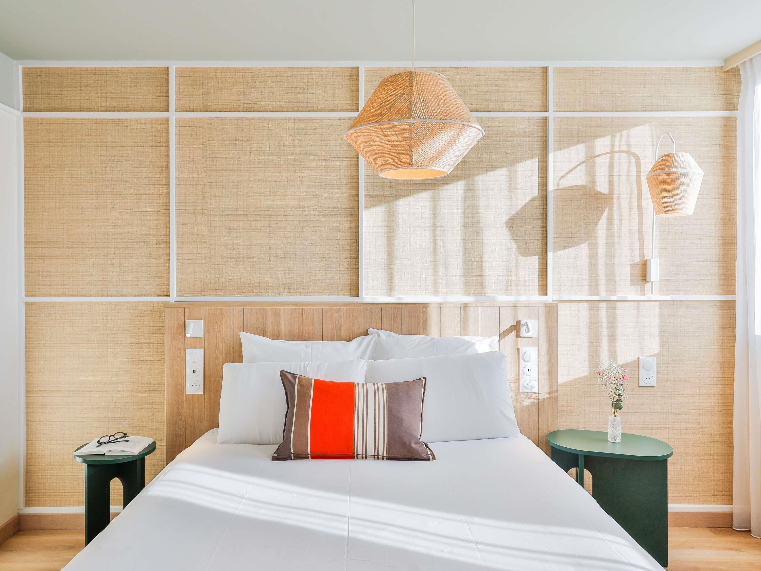 Une chambre d’hôtel moderne avec un grand lit, une table de chevet, une petite table de nuit verte et une lampe fixée au mur.