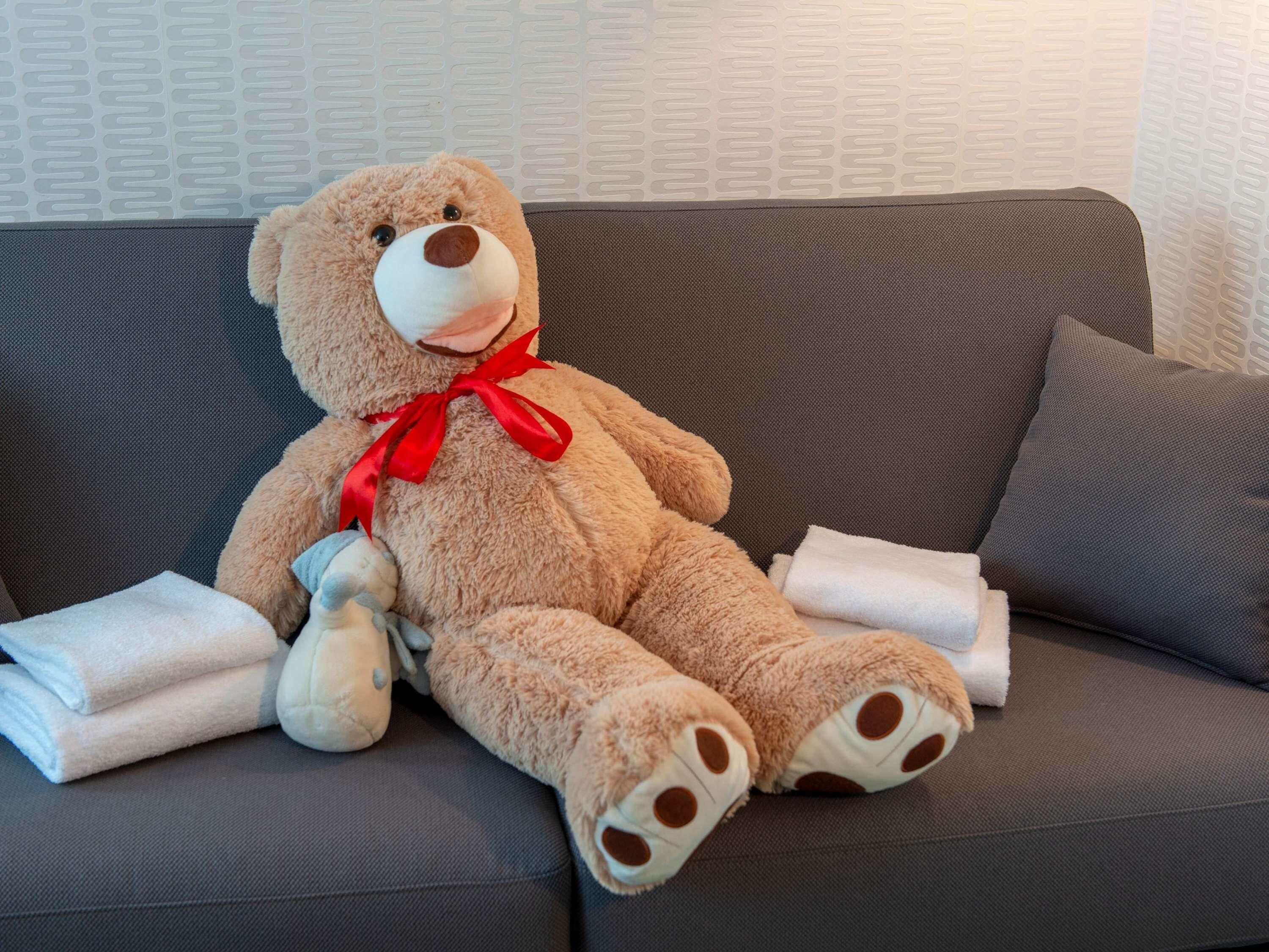 Un grand ours en peluche avec un nœud rouge est assis sur un canapé gris, et des serviettes blanches pliées sont posées à côté.