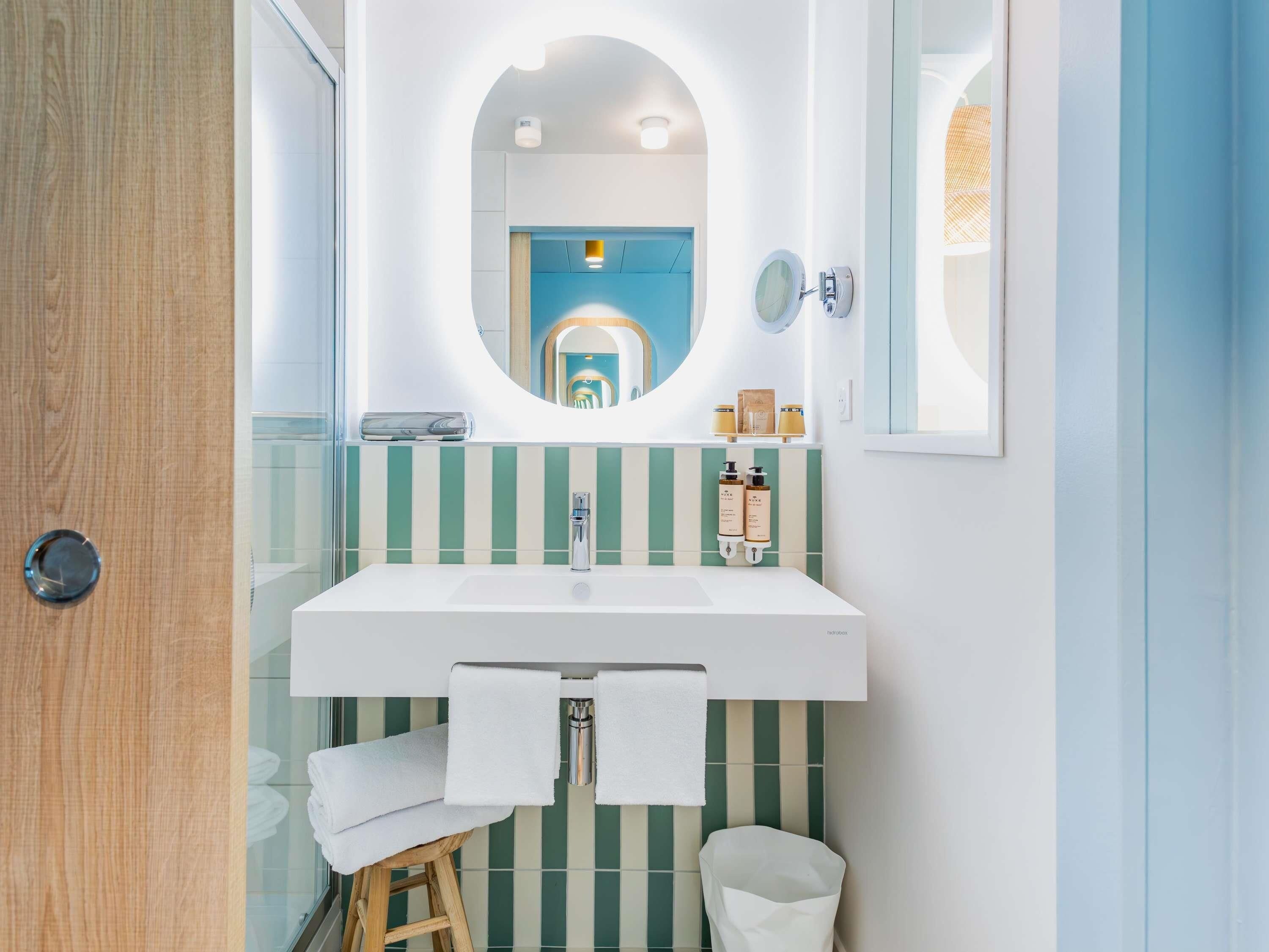 Une salle de bain avec un lavabo blanc, des carreaux à rayures vertes, un tabouret en bois et un miroir avec un cadre rond.
