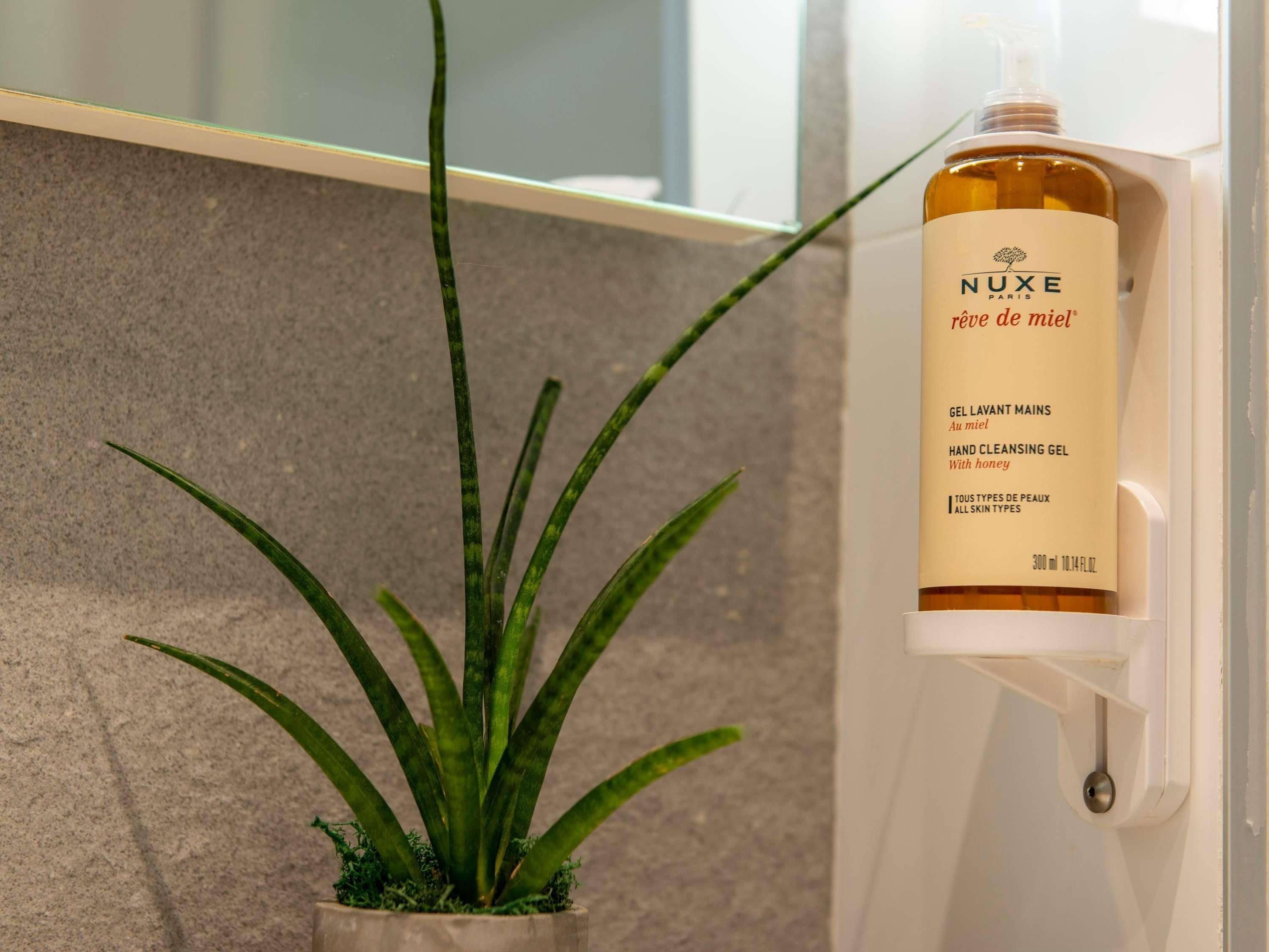 Una botella de gel limpiador de manos etiquetada 'NUXE PARIS' con dosificador de bomba, sobre una superficie blanca junto a una planta en maceta.
