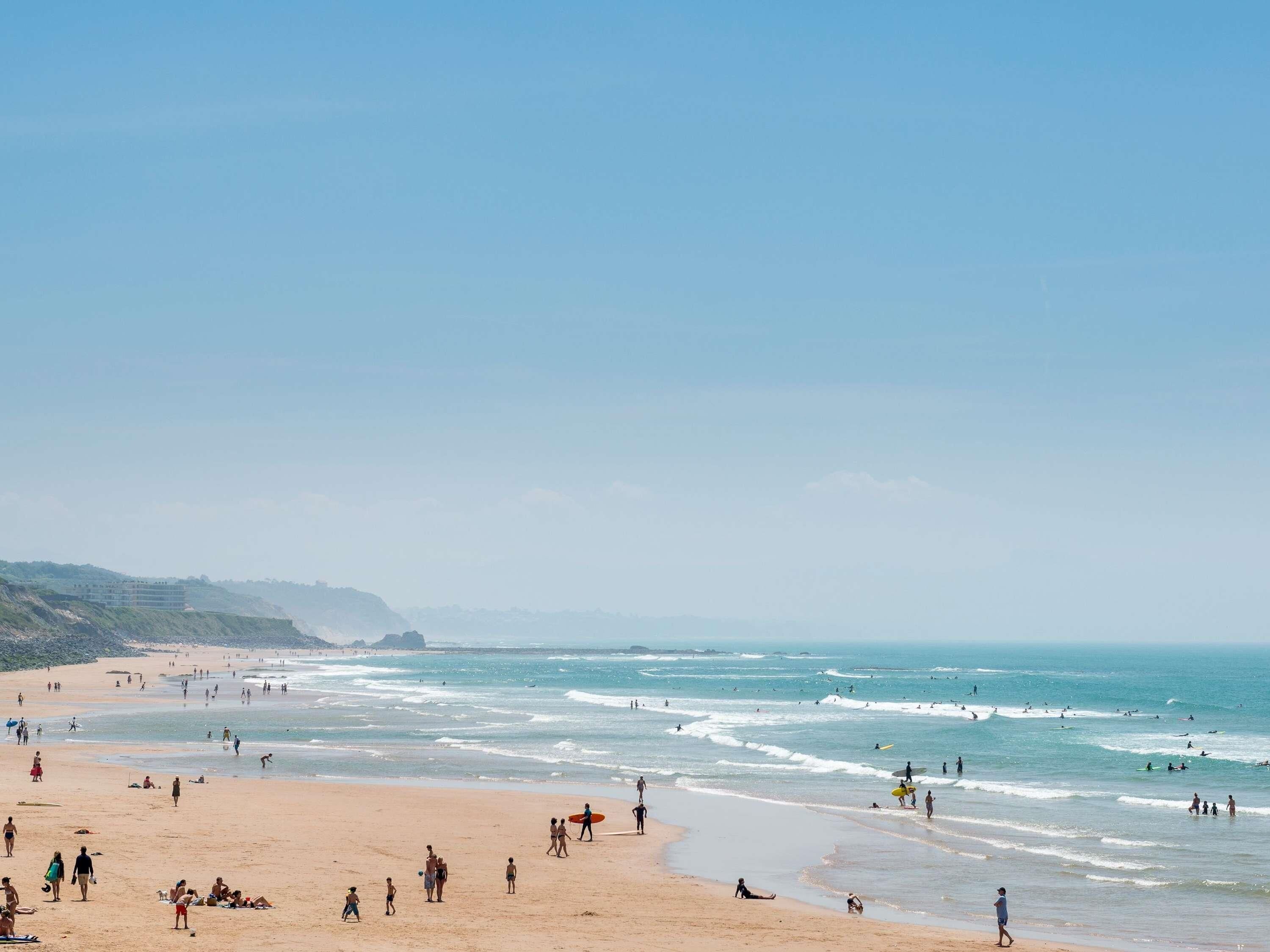 A beach with people walking and surfing, and a building in the background.