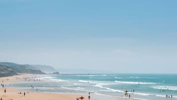 Una playa con gente caminando y practicando surf, y un edificio al fondo.