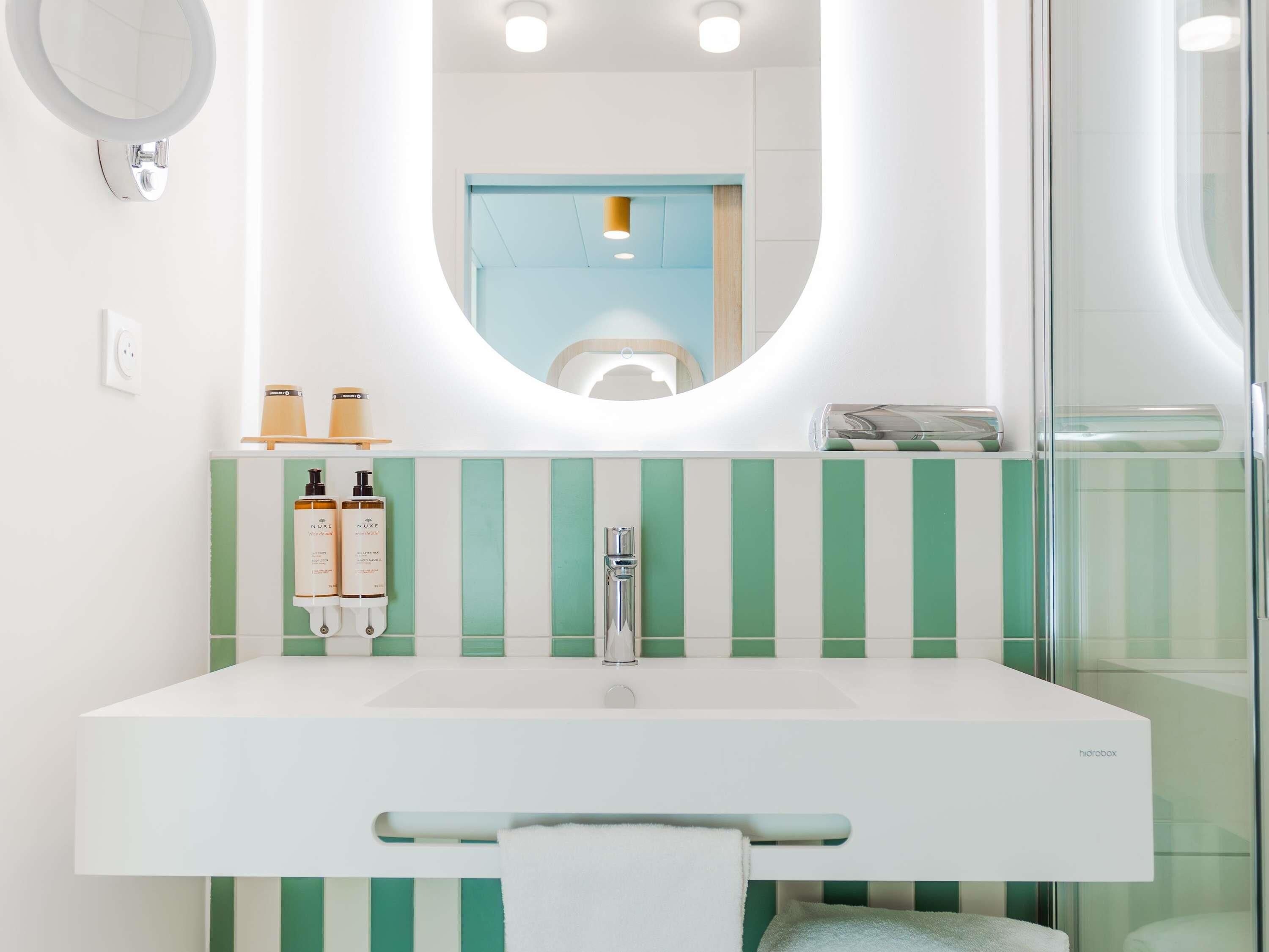 Un baño moderno con un lavabo blanco, baldosas verde a rayas, un espejo redondo y una cabina de ducha de cristal.