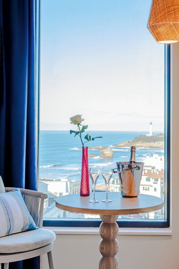 Una habitación con vistas al océano y un faro, una mesa con un florero y champán, y una silla con un cojín.