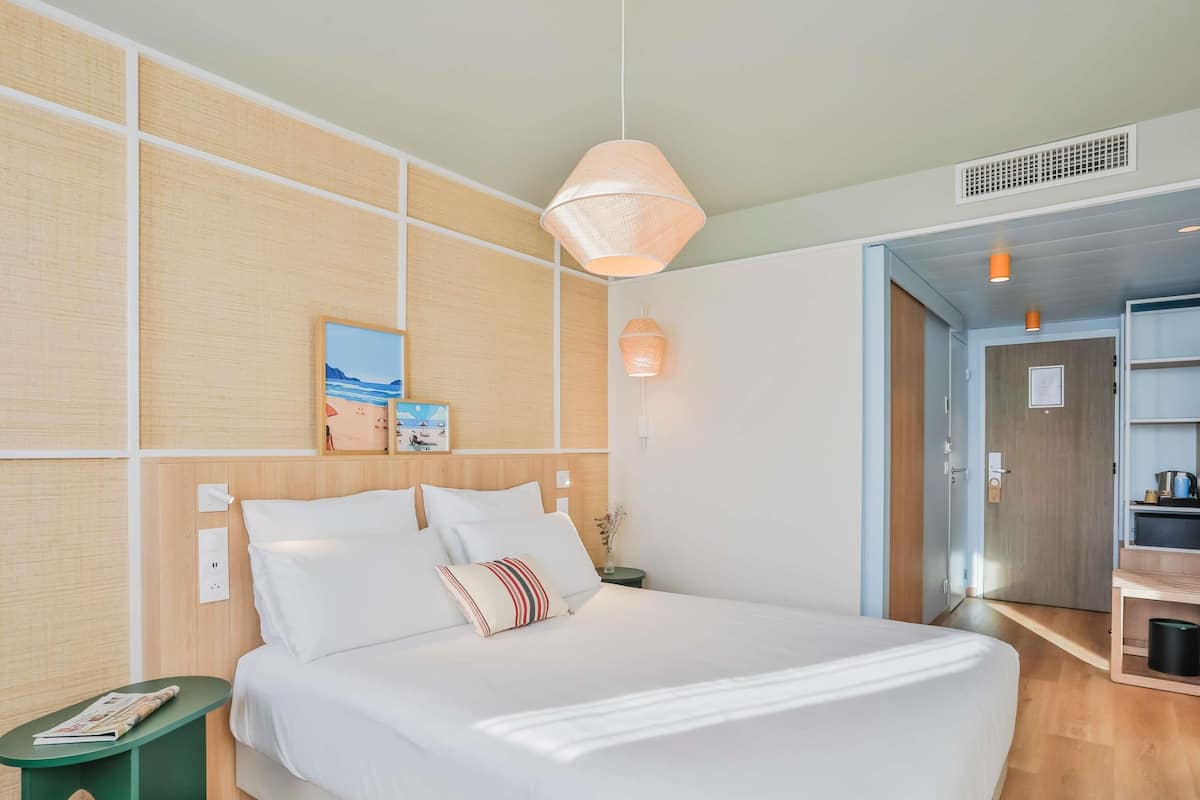 A neatly arranged hotel room with a large bed, a bedside table, a wall-mounted shelf, and a door leading to another room.
