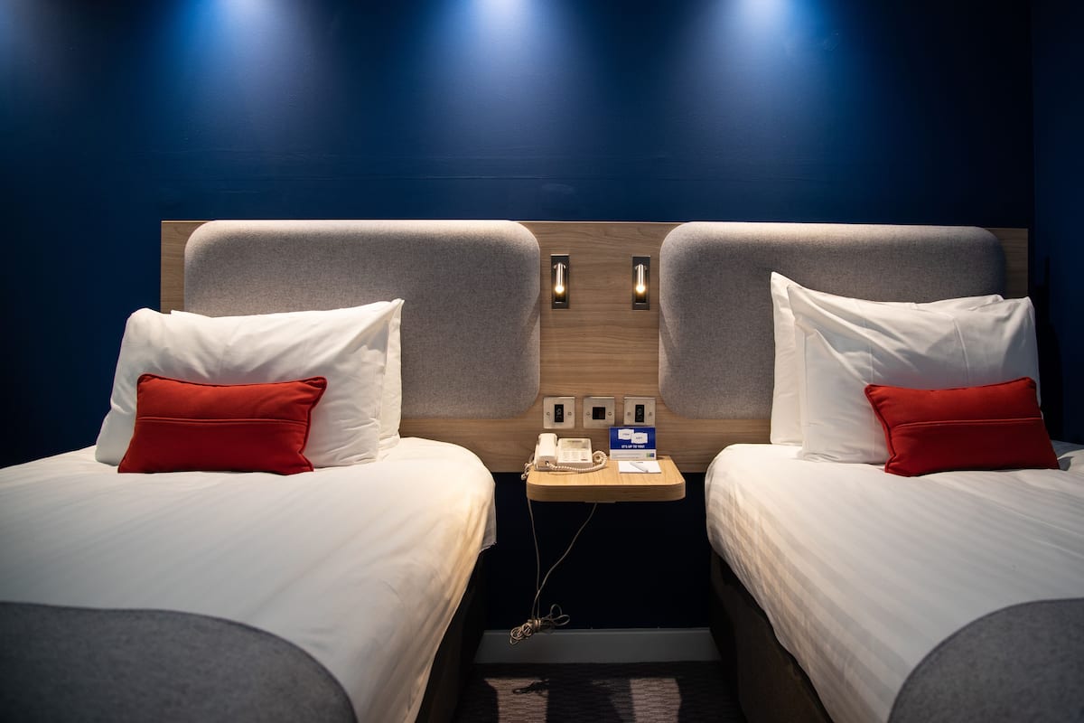 Hotelkamer met twee bedden, elk met witte lakens en rode kussens, een houten hoofdbord, een klein tafeltje met een telefoon en een bord, en twee wandlampen.