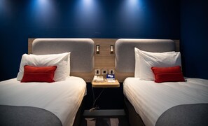 A hotel room with two beds, each with white bedding and red pillows, a wooden headboard, a small table with a telephone and a sign, and two wall-mounted light fixtures. - Holiday Inn Express London Stansted Airport by IHG (Bishop's Stortford)
