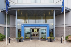 Entrada de un edificio moderno con toldo azul, puertas de vidrio y plantas en macetas.