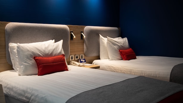 A hotel room with two beds, each with white bedding and red pillows. There is a wooden headboard, a bedside table with a lamp, and a wall-mounted phone.
