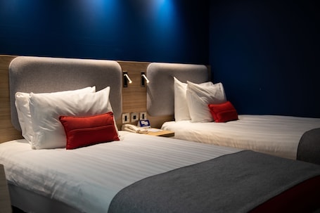 A hotel room with two beds, each with white bedding and red pillows. There is a wooden headboard, a bedside table with a lamp, and a wall-mounted phone.